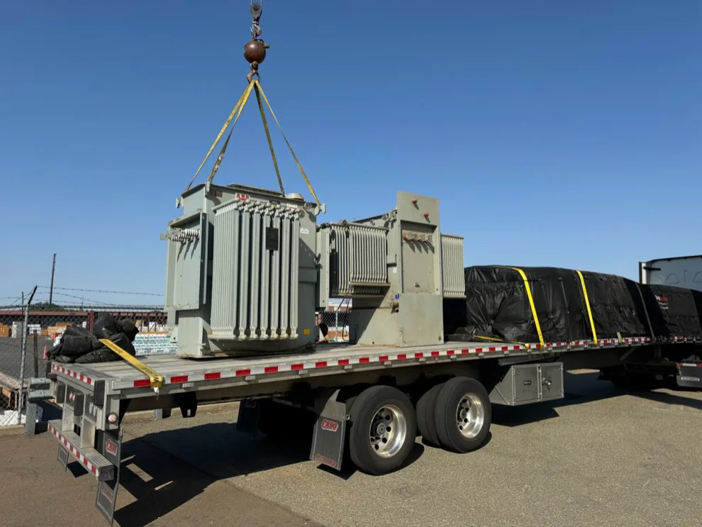 Electrical transformers being loaded onto a flatbed trailer with a crane.